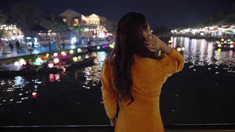 Behind-shot-of-Asian-woman-in-yellow-outfit-looking-at-night-river-reflections-in-Hoi-An,-Vietnam