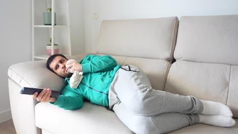 Man-with-toothache-lying-on-the-couch-while-turns-on-the-tv-and-holds-cheek-in-pain