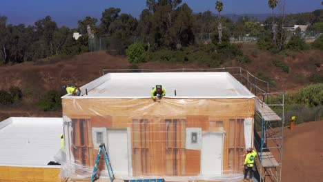 Close-up-descending-aerial-shot-of-a-construction-crew-working-on-the-siding-of-a-pre-fabricated-housing-module-at-a-building-site-in-West-Los-Angeles,-California