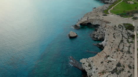 Eine-Luftaufnahme-Eines-Natürlichen-Felsbogens-über-Dem-Ruhigen-Blauen-Wasser-Des-Mittelmeers-In-Der-Nähe-Von-Ayia-Napa,-Mit-Einer-Zerklüfteten-Küste-Und-Einer-Grasfläche,-Die-Zum-Bogen-Hinaufführt