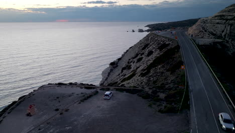 Vista-Aérea-Al-Atardecer-De-Una-Carretera-Costera-Cerca-De-La-Roca-De-Afrodita-En-Paphos,-Con-Un-Coche-Aparcado-Y-El-Tranquilo-Mar-Que-Se-Extiende-Hasta-El-Horizonte