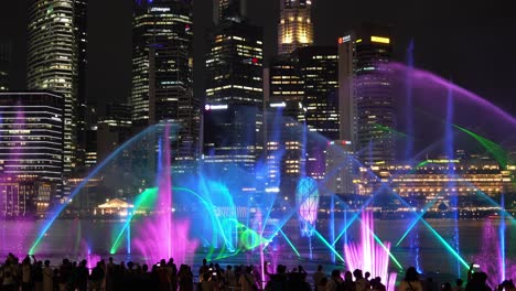 Crowd-of-audiences-at-the-Marina-Bay-Sands-event-plaza,-experiencing-a-magical-evening-with-SPECTRA-light-and-water-show,-glass-prism-in-the-center-and-downtown-cityscape-in-the-background,-timelapse