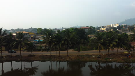 Toma-Aérea-De-Seguimiento-De-Palmeras-Y-El-Paisaje-Urbano-De-Ebolowa-Desde-Un-Lago-En-Camerún.