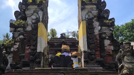 Mujeres-Bali-Con-Cestas-En-La-Cabeza-Atravesando-La-Puerta-Del-Antiguo-Templo-Hindú,-Ubud