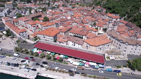 Kotor,-Montenegro,-Aerial-View-of-Old-Town-Buildings-and-Walls,-Traffic-on-Coastal-Road-and-Boast-in-Harbor,-Revealing-Drone-Shot