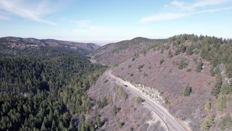 Cars-drive-along-a-road-on-a-mountain-surrounded-by-pine-tree-forest,-drone-aerial