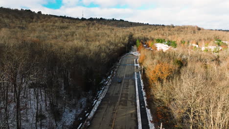 Vehículo-Conduciendo-Por-Una-Carretera-Vacía-Por-Un-Bosque-Invernal-Sin-Hojas,-Soleado,-Toma-Aérea