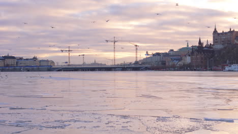 Twilight-drone-pullback-over-frozen-Riddarfjärden,-view-of-cranes-over-Slussbron