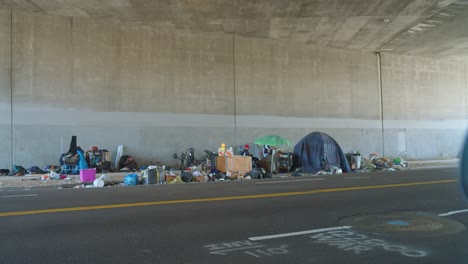 Homeless-encampment-under-an-overpass-with-personal-belongings-scattered-around,-daylight