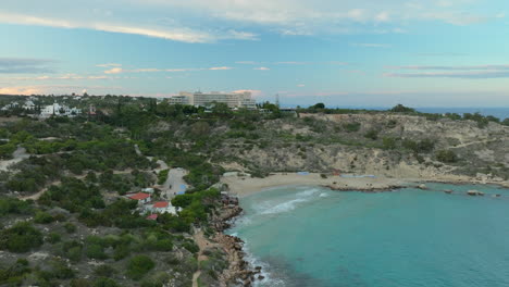 A-serene-aerial-view-of-Konnos-Beach-in-Cyprus,-featuring-a-secluded-sandy-cove-with-crystal-clear-turquoise-waters,-surrounded-by-lush-greenery-and-rugged-cliffs,-large-hotel-overlooking-the-bay
