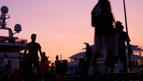 Zeitlupen-Nahaufnahme-Der-Spielersilhouetten-Während-Eines-Straßenfußballturniers-Bei-Sonnenuntergang-Mit-Yachten---Zuschauer-Im-Hintergrund