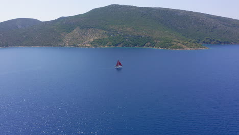 Aerial:-Slow-orbit-drone-shot-of-a-sailboat-with-red-sails-that-is-sailing-offshore-on-a-clear-summer-day