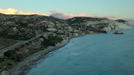 Dusk-view-of-a-winding-road-that-cuts-through-a-hilly-landscape-leading-to-a-coastal-beach-with-white-chalk-cliffs,-with-a-backdrop-of-pink-tinged-clouds-in-the-sky,-Pafos,-Afrodite-hills
