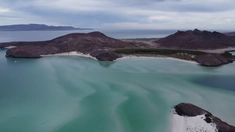 Playa-balandra-with-turquoise-waters-and-mountainous-backdrop,-baja-california,-mexico,-on-a-cloudy-day,-aerial-view