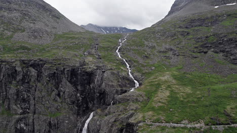 Wide-aerial-dolly-shot,-pushing-in-slowly-on-a-long,-thin,-rushing-river,-winding-down-a-steep,-mossy-hillside-by-Trollstigen,-Norway