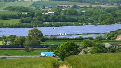 Un-Tren-Desde-La-Ciudad-De-Exeter-Y-El-Tráfico-De-La-Autopista-En-La-M5-Pasan-Entre-Un-Campo-De-Cebada-Y-Una-Gran-Granja-Solar-En-La-Exuberante-Campiña-De-Devon,-Cerca-De-La-Estación-De-Tren-Tiverton-Parkway.