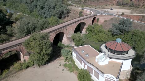 A-drone-captures-the-spa-building-and-its-lush-gardens,-with-the-backdrop-featuring-a-16th-century-bridge-spanning-the-Guadalquivir-River---all-part-of-the-Marmolejo-Spa-Center,-Andalusia,-Spain