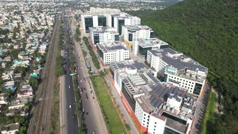 Aerial-Shot-of-Corporate-Buildings-near-a-hill-filled-with-trees-and-highway-filled-with-cars-passing-through