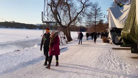 People-walk-and-run-on-snow-by-icy-river-in-sunny-Stockholm,-Sweden