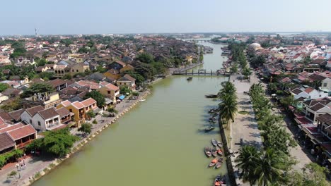 Drone-aerial-view-in-Vietnam-flying-over-Hoi-An-brown-color-river-canal-in-the-city,-small-brick-houses-and-palm-trees-on-a-sunny-day