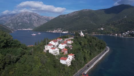 Kotor-Bay,-Montenegro,-Drone-Shot-of-Buildings-and-Church-on-Cape-Above-Sea-and-Ferry-Line-Boats-in-Kamenari