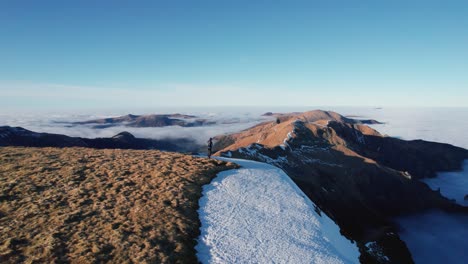 A-person-stands-on-a-mountain-ridge,-surrounded-by-snow-and-clouds-below,-in-the-Massif-Central-Auvergne-Volcanoes-National-Park-in-France