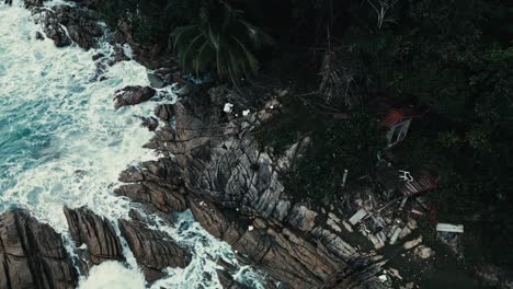 Aerial-orbit-shot-of-small-hut-on-rocky-coastline-cliff-with-foamy-white-waves-splashing-ashore