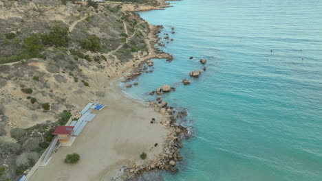 Aerial-image-captures-a-gentle-arc-of-sandy-beach-tucked-between-rugged-cliffs-and-the-azure-waters-of-the-Mediterranean,-with-a-smattering-of-boulders-along-the-shoreline-and-beach-facilities-nearby