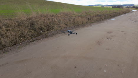 The-drone-hovers-above-a-muddy-path-surrounded-by-tall,-dried-grass,-with-fields-and-a-hilly-terrain-in-the-distance