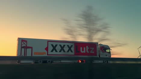 XXXLUTZ-delivery-truck-drives-on-motorway-in-front-of-wind-turbines-at-sunset