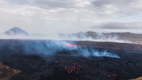 Lava-flows-at-Fagradalsfjall,-smoky-scene-with-mountains-in-backdrop,-daylight