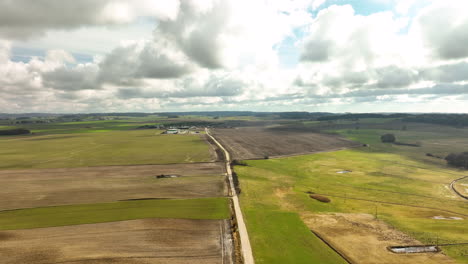 An-aerial-view-captures-a-sweeping-landscape-of-agricultural-fields-in-Poland,-with-varying-hues-of-green-and-brown-under-a-dramatic-sky-dotted-with-clouds,-road-that-cuts-through-it