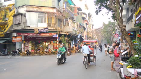 Bustling-street-scene-in-Hanoi-at-dusk-with-cyclos-and-motorbikes,-local-shops-lining-the-road