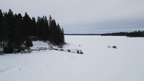 Eine-Langsame-Drohnen-Landschaftsaufnahme-Einer-Insel-Auf-Dem-Zugefrorenen-Kanadischen-Paint-Lake-Mit-Einer-Eisfischerhütte-Und-Skioos