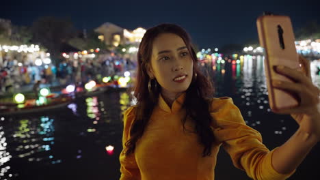 Una-Atractiva-Mujer-Vietnamita-Con-Un-Tradicional-Vestido-Amarillo-Se-Toma-Una-Foto-Selfie-Nocturna-Junto-Al-Río-En-Hoi-An,-Vietnam