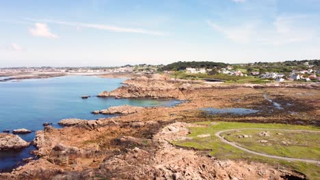 Guernsey-shoreline-flying-over-headland,bays,rocks-and-clear-blue-sea-with-beaches,-fields-and-houses-in-the-distance-on-bright-sunny-day