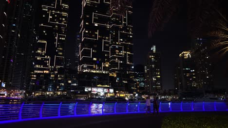 Dubai-Marina-at-Night,-People-Walking-on-Promenade-by-Water-and-Skyscrapers-in-Lights