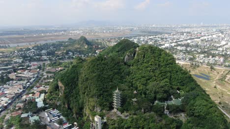 Drone-aerial-view-in-Vietnam-flying-over-Da-Nang-city-marble-mountains-full-of-trees-with-buddhist-temples-surrounded-by-flat-land-on-a-sunny-day