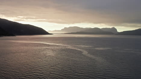 Aerial-view,-climbing-over-Sognefjord-in-Norway,-moving-toward-a-sunset-over-distant-shadowy-mountains-and-islands