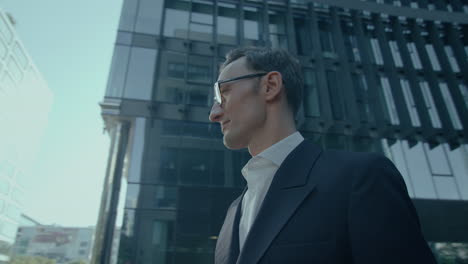 Elegant-Handsome-Caucasian-Man-In-Suit-and-Glasses-Outdoors-Looking-Aside-At-Camera,-Orbital-Low-Angle-Shot