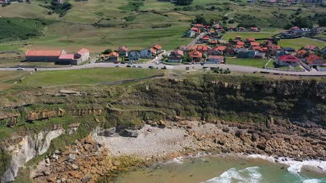 Toma-Aérea-En-La-Costa-Del-Mar-Cantábrico-Hacia-El-Acantilado-Hay-Una-Carretera-Por-La-Que-Pasan-Los-Coches-Junto-A-Casas-Típicas-En-Prados-Verdes-Con-El-Cielo-Al-Fondo-Y-Nubes-En-Cantabria-España