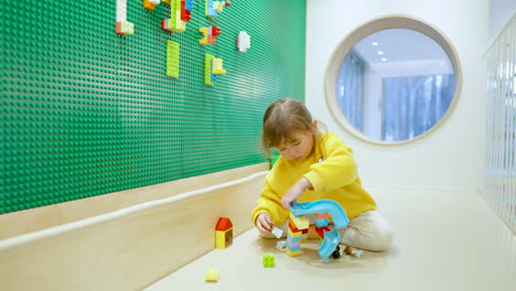 Little-Adorable-Cute-Caucasian-American-Girl-Playing-With-Colorful-Lego-Blocks-Building-Ramp-Bringe-At-Indoor-Playroom-Sitting-on-a-Floor---slow-motion-wide