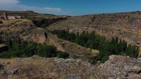 flight-with-a-surprise-effect-watching-the-course-of-a-canyon-and-discovering-a-beautiful-town-with-its-tower-and-its-dry-meadows-in-summer-with-a-blue-sky-in-Segovia-Spain