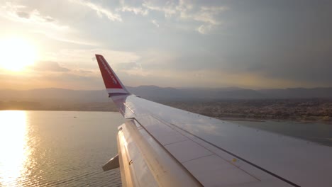 Wing-of-Norwegian-airplane-before-landing-by-coast-at-sunset,-slomo