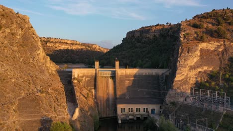 Drone-flight-ascending-in-front-of-a-hydroelectric-plant-on-a-water-dam-built-on-the-edges-of-an-orange-stone-canyon-with-forest-formations-of-a-river-at-the-golden-hour-in-Segovia-Spain
