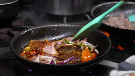 Chef-Profesional-De-Restaurante-Voltea-Con-Gracia-Verduras-Salteadas-En-Una-Sartén-Chisporroteante-Sobre-Una-Llama-De-Alto-Calor-En-La-Estufa,-Las-Chispas-Vuelan-Mientras-El-Cocinero-Sacude-La-Sartén-Sobre-El-Quemador-De-Gas-De-La-Cocina,-Cámara-Lenta-De-Cerca-4k