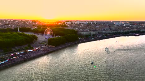 Wine-fair-with-illuminated-Ferris-wheel-at-sundown-with-crowds-in-Bordeaux-France-with-Garonne-river-and-boats,-Aerial-flyover-shot