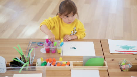 Una-Niña-Rubia-De-3-Años-Estudia-Pintura-Con-Cuentagotas-Sentada-Junto-Al-Escritorio,-Chupa-Agua-Coloreada-En-Una-Pipeta-De-Contenedores