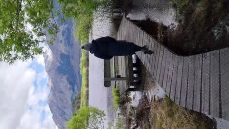 A-vertical-of-a-woman-walking-on-a-wooden-boardwalk-with-a-view-of-a-lake-and-snow-capped-mountains-in-Glenorchy-New-Zealand