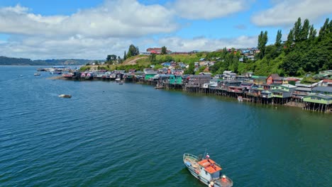 Colorful-castro-stilt-houses-on-chiloe-island-with-a-boat-cruising,-aerial-view